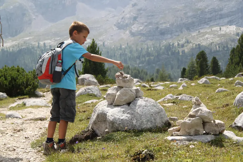 A boy with a backpack stacking stones into little towers on a rocky mountain meadow in the Dolomites