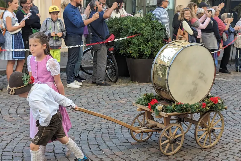 A girl and a boy in traditional dress pulling a decorated cart with a big drum at the Merano Grape Festival