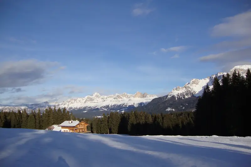 Eine Almhütte aus Holz am Rand einer verschneiten Wiese und eines Fichtenwalds, dahinter verschneite Dolomitengipfel