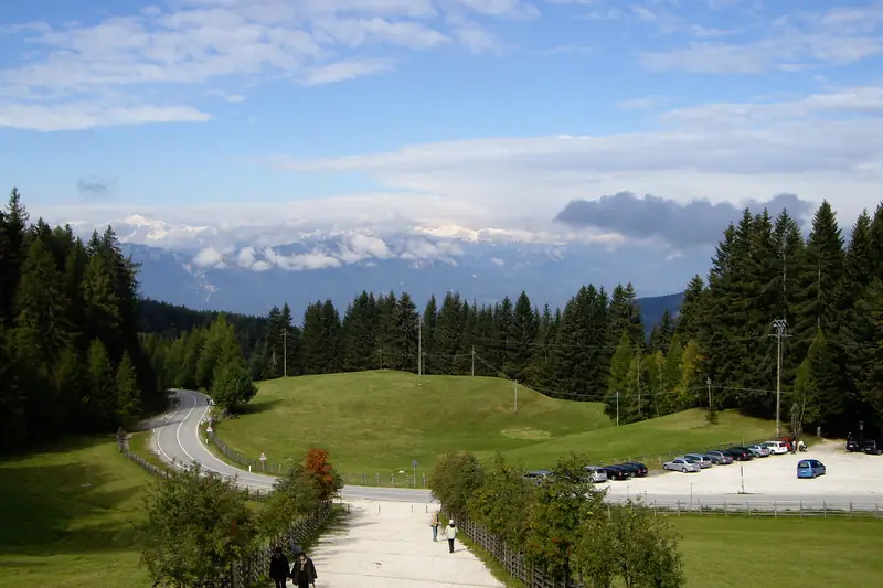 Ein Kiesweg und grüne Wiesen beim Parkplatz von Maria Weißenstein, dahinter Wald und Berge unter blauem Himmel